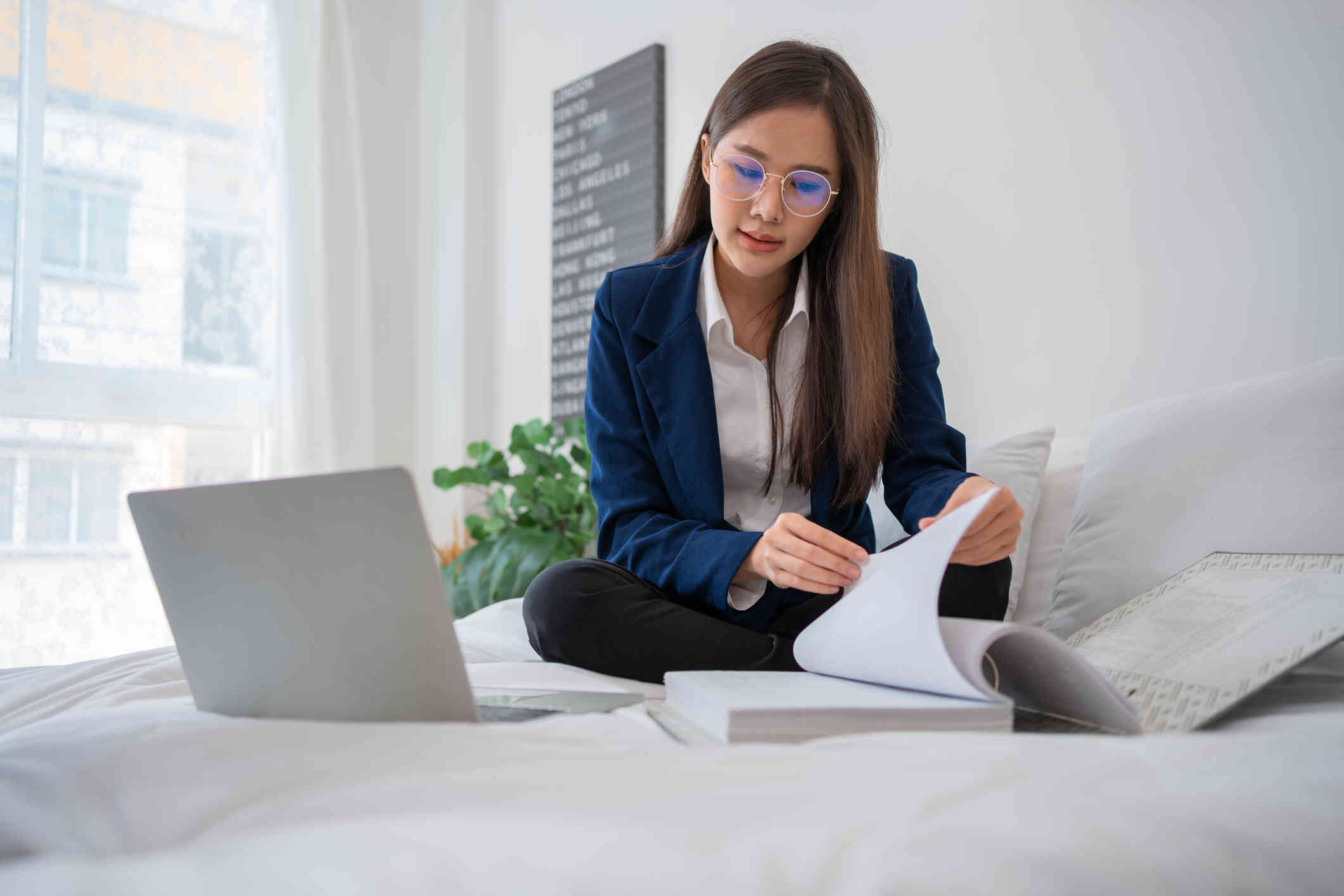 A woman in a blue bazer sits cross legged on the bed with her laptop while looking at a large book.